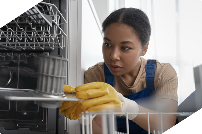 A technician inspects the inside of a dishwasher during a repair, wearing yellow gloves for safety.