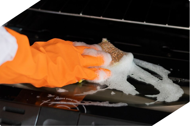 A close-up of a person scrubbing an oven door with soapy water, wearing bright orange cleaning gloves.