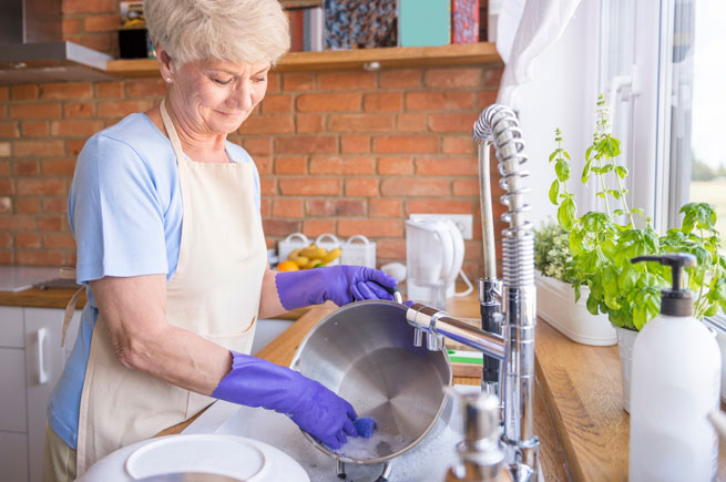 Elderly woman wearing gloves hand-washing dishes in a bright kitchen with a sunny window.