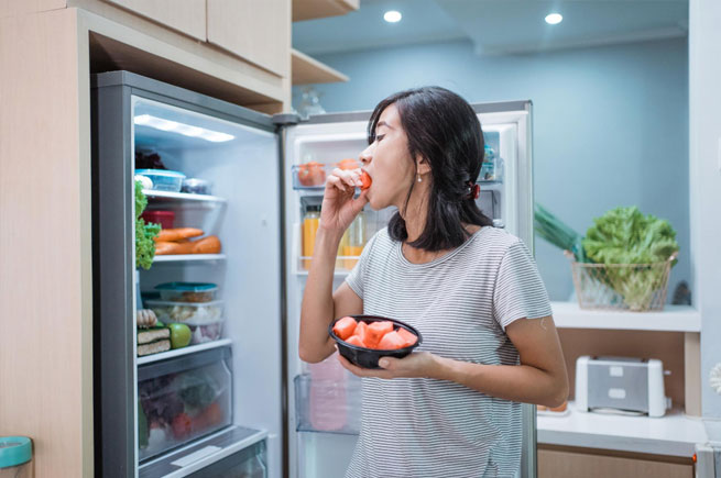 A woman eating fresh watermelon from the fridge, standing in a modern kitchen with a well-organized refrigerator.