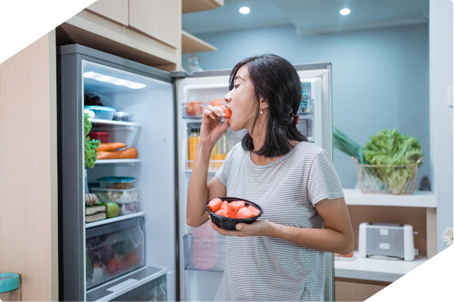 A woman eating fresh watermelon from the fridge, standing in a modern kitchen with a well-organized refrigerator.